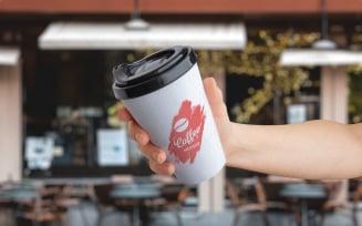 Logo mockup on a paper takeaway cup held in hand in front of a coffee shop