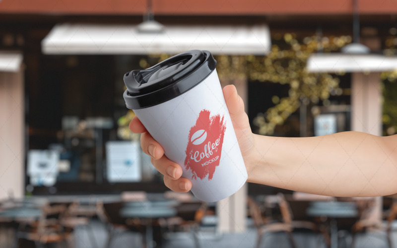 Maqueta del logotipo en un vaso de papel para llevar sostenido en la mano frente a una cafetería